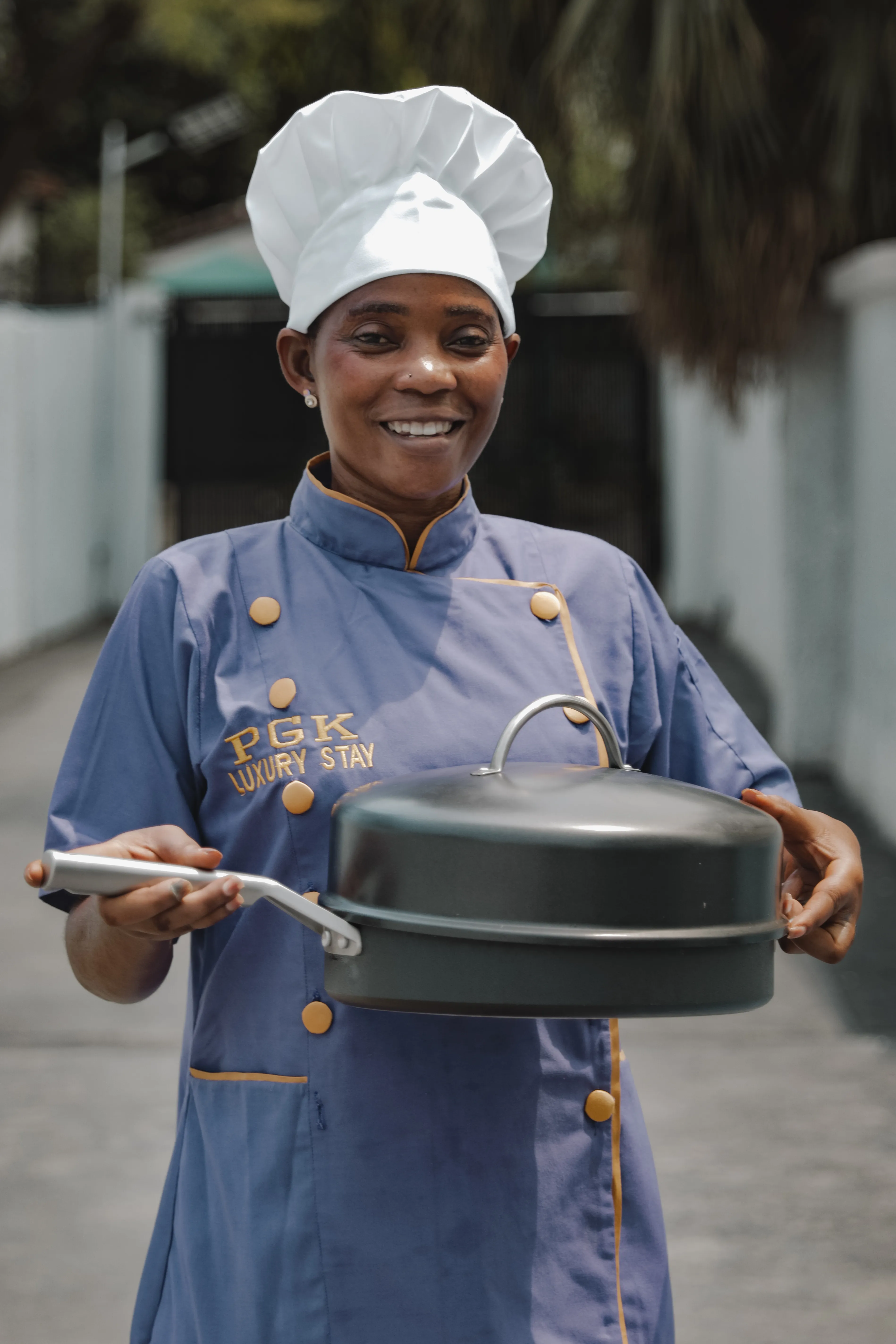 Smiling chef in a branded uniform and toque presenting a covered dish outdoors.