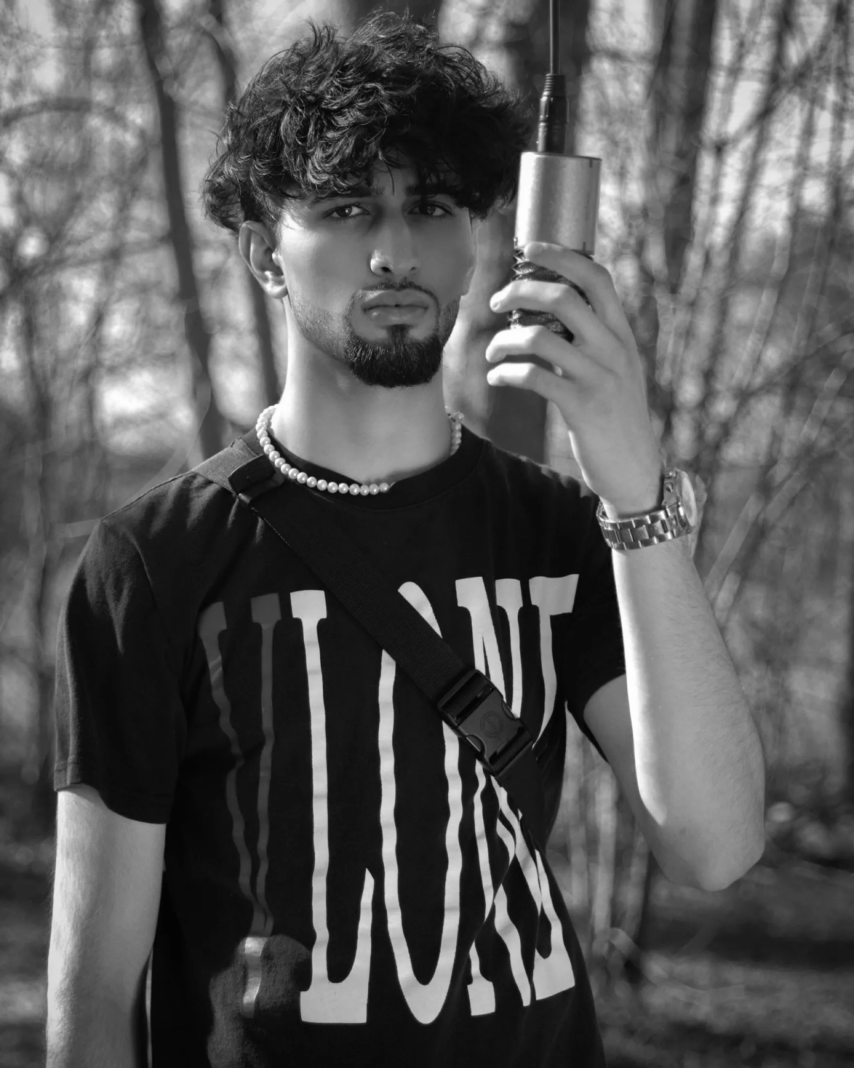 Black-and-white portrait of a man with a microphone, chain and graphic tee in woodland.