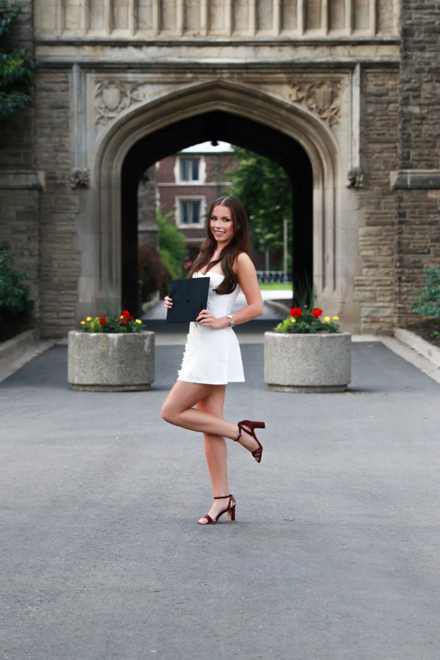 Graduate posing with her diploma in front of a campus archway and garden.