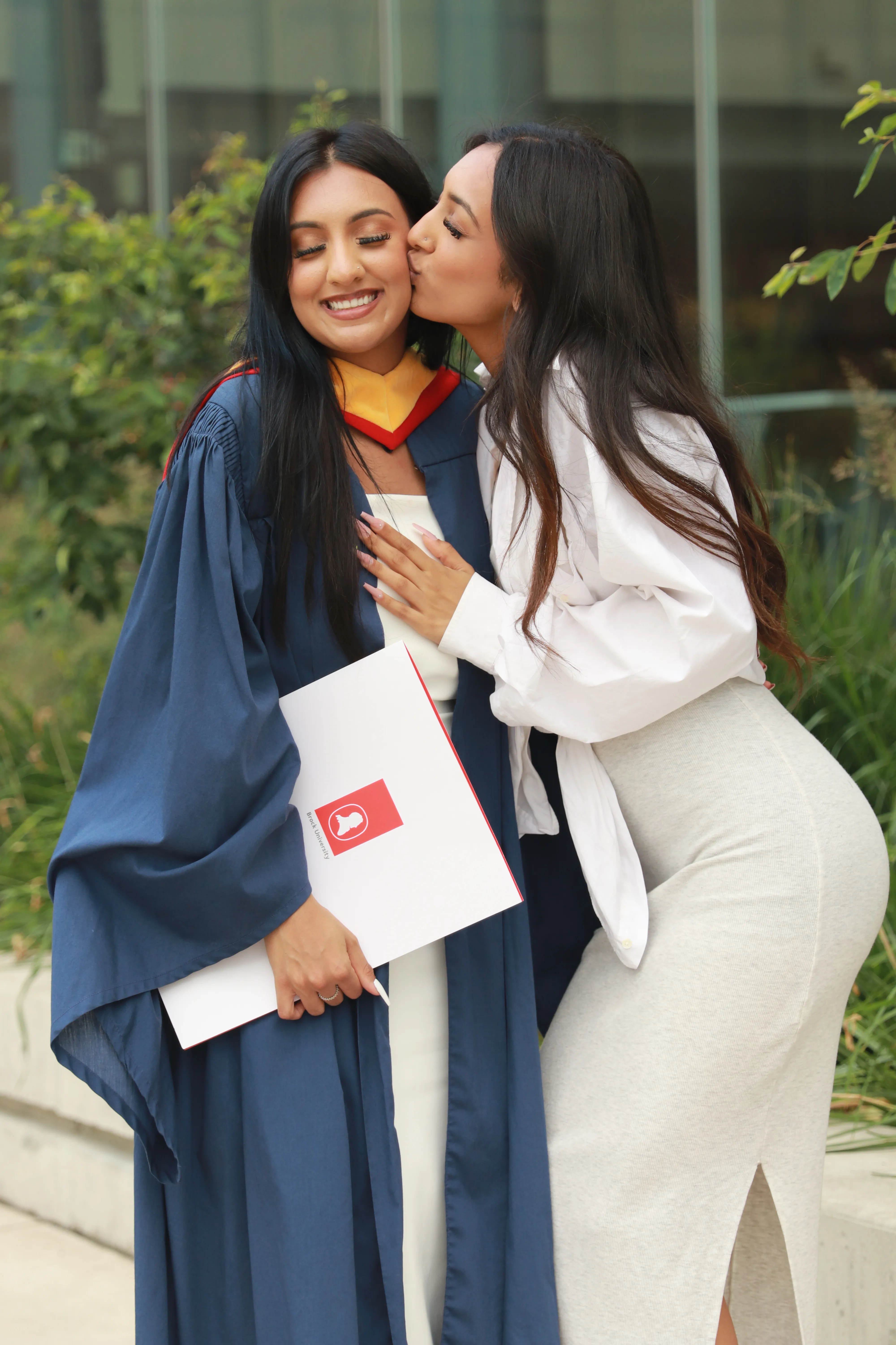 Friend kissing a graduate on the cheek outside a university building.