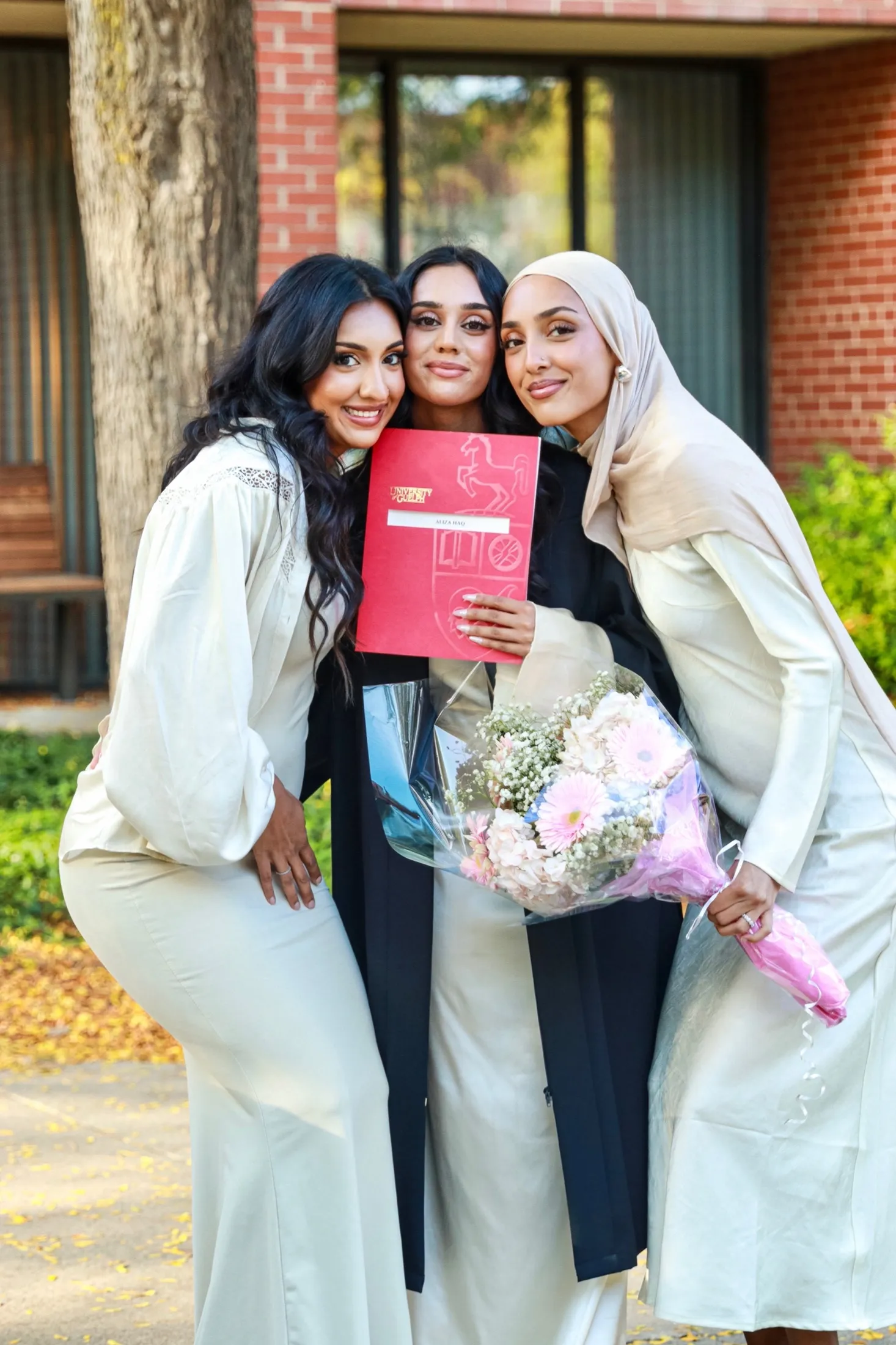 Graduate with two family members holding flowers and her diploma outdoors.