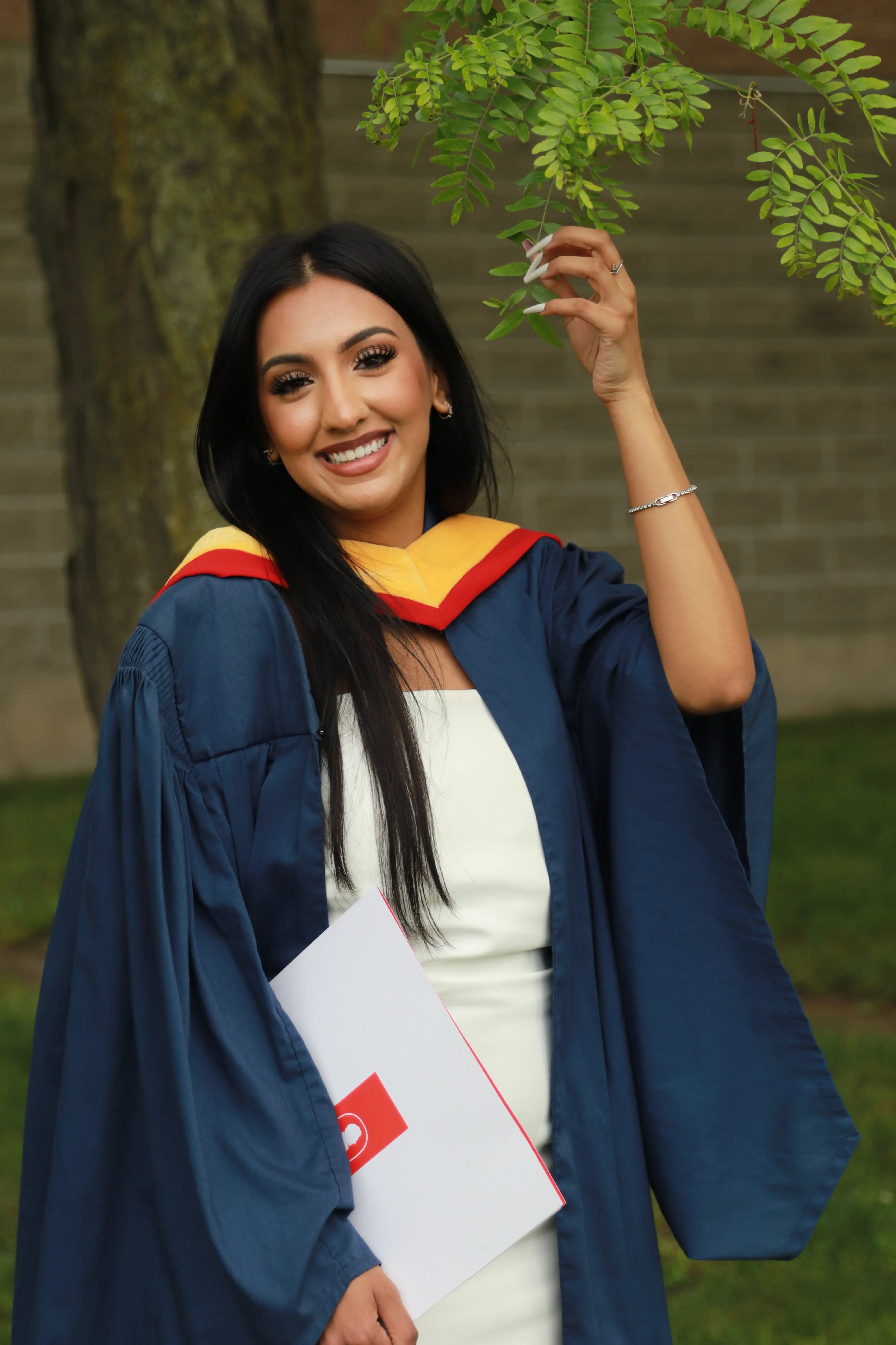 Graduate in cap and gown reaching up to a tree branch, holding her diploma.