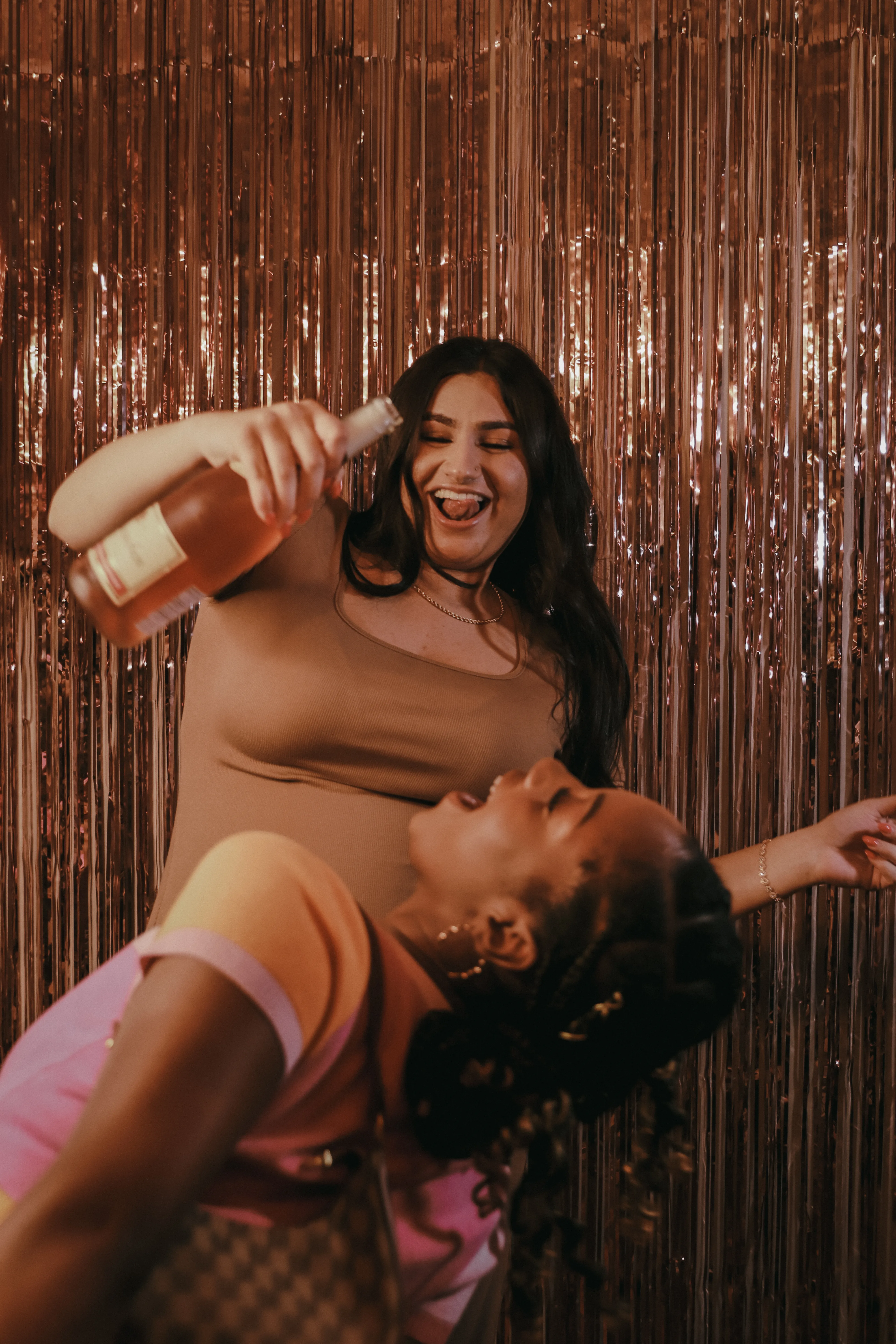 Two women laughing and dancing against a rose-gold fringe backdrop.
