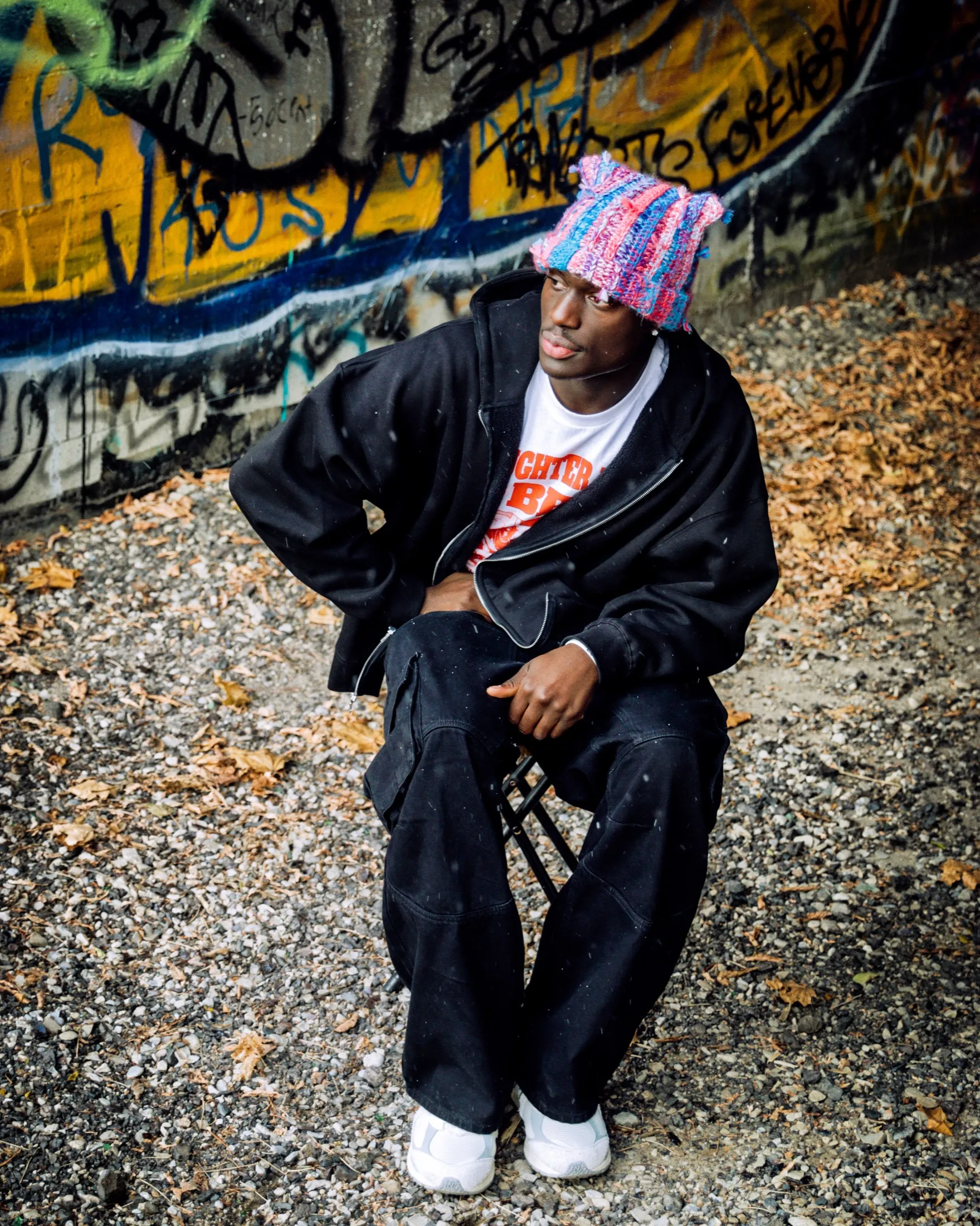 Man in a pink knit beanie and black streetwear seated against a graffiti wall.