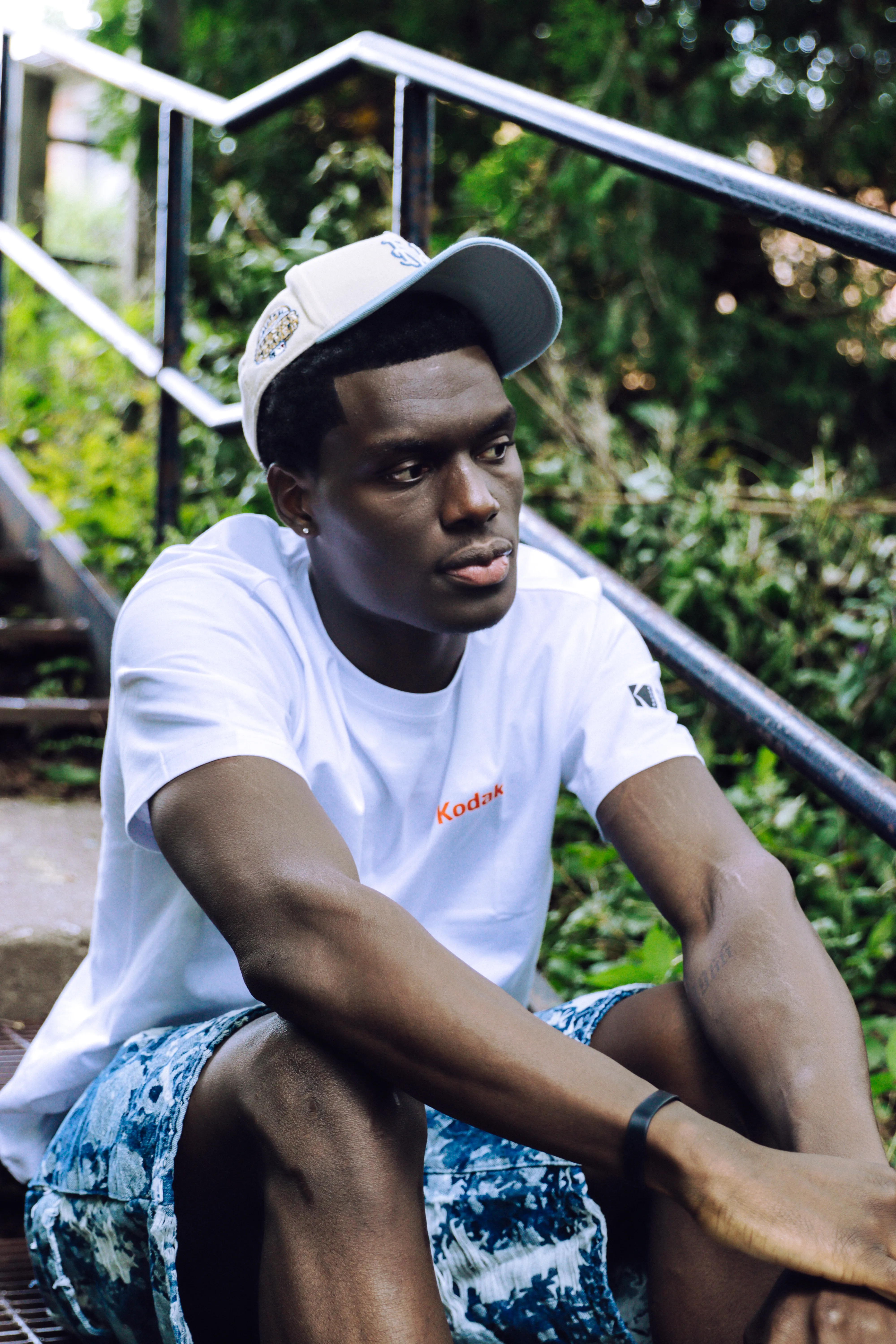 Man in a Kodak T-shirt and cap seated on outdoor stairs surrounded by greenery.